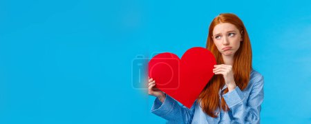 Photo for Gloomy girl was rejected on valentines day. Uneasy upset redhead female in nightwear, holding heart card and sobbing, look away upset, feeling depressed, standing blue background. - Royalty Free Image