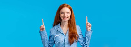 Photo for Charismatic and energized good-looking redhead female in cute pyjama pointing raised fingers up and smiling, invite for sleepover, introduce discount or promos, standing blue background. - Royalty Free Image
