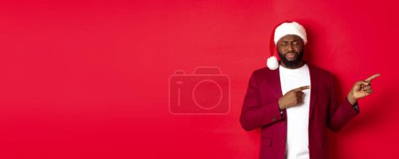 Téléchargez les photos : Noël, fête et concept de vacances. Homme afro-américain bouleversé regardant sombre, les yeux fermés et soupirant, les doigts pointant vers la droite, debout dans le chapeau de Père Noël sur fond rouge. - en image libre de droit