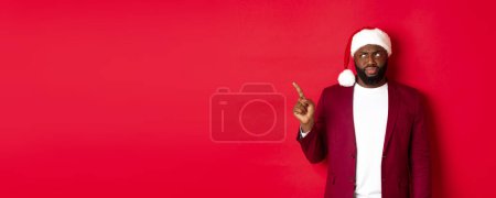 Foto de Concepto de Navidad, fiesta y vacaciones. Escéptico y sombrío hombre afroamericano apuntando con el dedo hacia la izquierda, mirando decepcionado, de pie en sombrero de santa sobre fondo rojo. - Imagen libre de derechos