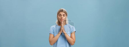 Photo for Lifestyle. Good-looking kind and faithful european girl with blond hair smiling cute and tender holding hands in pray while making wish hopefully believing god hearing her prayers with closed eyes - Royalty Free Image