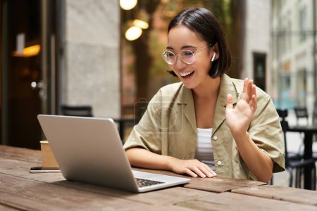 Photo for Portrait of young woman video chatting, having online meeting on laptop, sitting in outdoor cafe with compute, wearing glasses and smiling. - Royalty Free Image