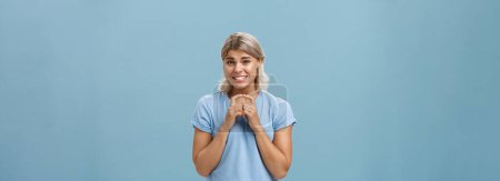 Photo for Silly cute and worried blonde female friend with short haircut steepling index fingers and smiling with guilty sorry smile frowning while waiting for answer begging for help or favor over blue wall - Royalty Free Image