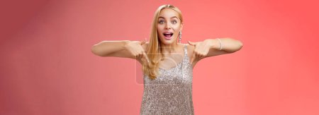 Photo for Impressed excited attractive glamour blond girl in silver glittering dress gasping thrilled pointing down glance camera fascinated check out fabulous awesome promo, standing amused red background. - Royalty Free Image