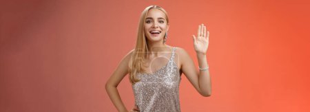 Photo for Lifestyle. Friendly stylish glamour attractive blond caucasian woman in silver glittering dress hold hand waist waiving palm hello hi what up gesture smiling introducing herself party boyfriend friend - Royalty Free Image