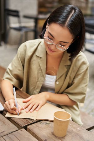 Foto de Tiro vertical de una joven asiática haciendo deberes, tomando notas, escribiendo algo, sentada en un café al aire libre y bebiendo café. - Imagen libre de derechos