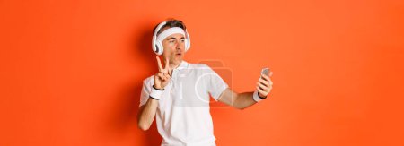 Photo for Portrait of attractive middle-aged male athlete, taking selfie during workout training, showing peace sign, listening music in headphones, standing over orange background. - Royalty Free Image