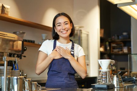 Foto de Amistosa chica asiática sonriente barista, toma de la mano en el corazón, mira agradecido al cliente, ama a sus invitados en la cafetería, se para en delantal uniforme. - Imagen libre de derechos