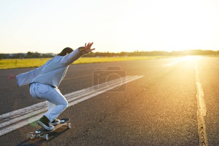 Téléchargez les photos : Joyeux patineur fille chevauchant son skateboard et s'amusant dans la rue vide. Femme souriante appréciant croisière sur route ensoleillée. - en image libre de droit