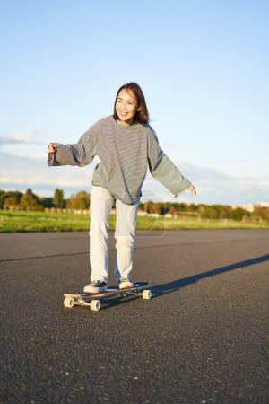 Téléchargez les photos : Mignon asiatique fille équitation skateboard, patinage sur la route et souriant. Skater sur le longboard cruiser profiter en plein air par une journée ensoleillée. - en image libre de droit