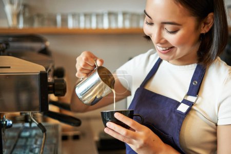 Smiling asian barista girl, making coffee, pouring steamed milk into cappuccino, doing latte art in cup, working in cafe.