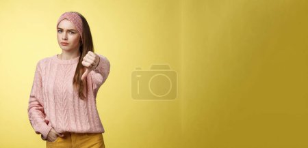 Photo for Not cool. Frustrated displeased and disagree cute young woman wearing knitted sweater frowning showing thumb-down smirking judgemental, expressing disapproval of unwell situation over yellow wall. - Royalty Free Image