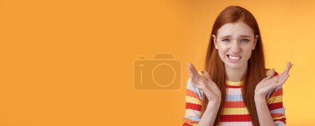 Photo for Awkward unhappy worried young redhead girl cringe feel sorry apologizing smirking smiling nervously frowning squinting spread hands sideways shrugging confused, standing orange background. - Royalty Free Image
