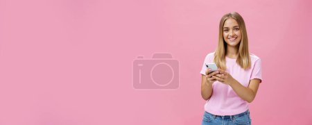 Téléchargez les photos : Femme appelant taxy via smartphone demandant l'adresse d'un ami souriant joyeusement à la caméra tenant le téléphone avec les deux mains sur la poitrine entrer en contact avec les clients via des messages debout sur le mur rose - en image libre de droit