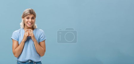 Photo for Silly cute and worried blonde female friend with short haircut steepling index fingers and smiling with guilty sorry smile frowning while waiting for answer begging for help or favor over blue wall - Royalty Free Image