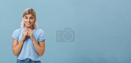 Photo for Lifestyle. Studio shot of insecure and timid silly girlfriend with blond hair clenching teeth looking shy down and steepling index fingers unconfident trying ask out boy she likes over blue background - Royalty Free Image