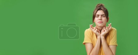 Photo for Worried intense, concerned attractive young european female model in yellow t-shirt raising shoulders crossing fingers for good luck pursing lips and closing eyes while waiting for miracle hopefully - Royalty Free Image