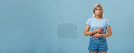 Téléchargez les photos : Portrait de jeune fille blonde intense mécontente avec des tatouages fronçant les sourcils touchant l'estomac se plaignant de maux d'estomac après avoir mangé de la malbouffe ayant un trouble sur fond bleu. Santé - en image libre de droit