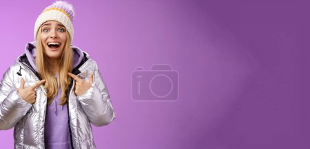 Téléchargez les photos : Surpris heureux souriant joyeux attrayant femme pointant elle-même satisfait choisi gagnant debout excité en argent brillant veste chapeau d'hiver ne peut pas croire sa propre chance, fond violet. - en image libre de droit