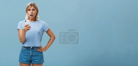 Photo for Worried confused young blonde woman in trendy outfit looking concerned and anxious realising bad thing happened receiving shocking news via smartphone opening mouth and staring at camera. Technology - Royalty Free Image
