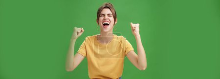 Foto de Foto cintura-up de mujer carismática energizada y excitada en camiseta amarilla cerrando los ojos gritando de alegría y felicidad levantando las manos en alegría celebrando noticias exitosas sobre fondo verde - Imagen libre de derechos