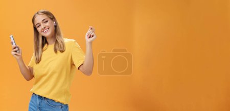 Foto de Mujer europea alegre y despreocupada que usa auriculares inalámbricos que sostienen el baile del teléfono inteligente con las manos levantadas y sonrisa feliz complacida que lleva vanlife tener tiempo para relajarse y disfrutar de la vida sobre la pared naranja - Imagen libre de derechos