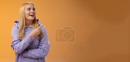 Amüsiert unbeschwerte fröhliche blonde Europäerin in Kapuzenpulli, die nach rechts zeigt, erfreut lächelnd glücklich interessante faszinierende Ausstrahlung genießt, die Auswahl im Laden trifft, orangefarbener Hintergrund.