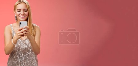 Téléchargez les photos : Délicieux tendre glamour femme blonde en argent élégant robe scintillante boucles d'oreilles brillant tenant smartphone prenant photo ami capture moment célébration boîte de nuit, fond rouge souriant. - en image libre de droit