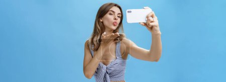 Téléchargez les photos : Femme glamour sortante et auto-assurée comme prendre des photos d'elle-même tenant smartphone faisant des lèvres pliantes selfie sensuelles et flirtantes levant la paume pour envoyer un baiser de vent aux adeptes aimants l'écran - en image libre de droit