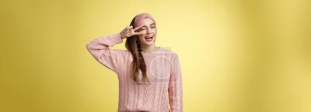 Foto de Alegre feliz glamour joven mujer europea en suéter de punto rosa, usando diadema, guiñando un ojo coqueto y lindo, mostrando la victoria o signo de paz sobre el ojo, sintiéndose emocionado y alegre sobre la pared amarilla. - Imagen libre de derechos