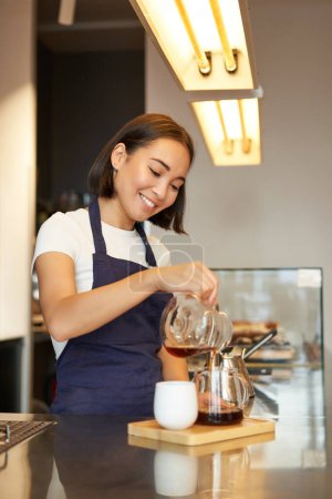 Téléchargez les photos : Plan vertical de fille souriante travaillant comme barista, préparer verser plus, fabrication de café filtre mélange, debout au comptoir dans le café dans le tablier. - en image libre de droit