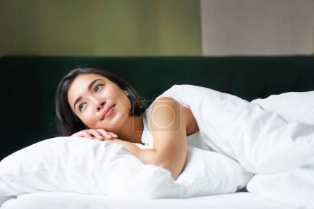 Foto de Soñadora chica asiática tumbada en su cama por la mañana, cubierta de tibio duver y apoyada en una almohada blanca, mirando a la ventana y pensando. - Imagen libre de derechos
