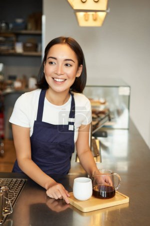 Téléchargez les photos : Plan vertical de fille souriante barista servant du café, fabrication de bière par lots, filtre pour le client dans le café, portant tablier bleu derrière le comptoir. - en image libre de droit