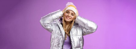 Photo for Tender romantic attractive blond female enjoying winter ski resort vacation having fun look pleased smiling broadly tilting head touching hat wearing silver stylish jacket, purple background. - Royalty Free Image