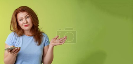 Photo for Well meh. Indifferent careless hesitant redhead middle-aged woman mature red female shrugging hold smartphone smirk bored uninterested hold hand aside apathetic attitude green background. - Royalty Free Image