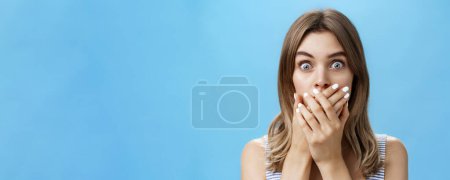 Photo for Close-up shot of shocked speechless woman covering mouth with pressed palms popping eyes at camera from surprise and worry, gasping witnessing terrible scene, feeling scared and intense over blue wall - Royalty Free Image