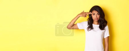 Photo for Portrait of skeptical and bothered african-american woman, making finger gun sign over head and smirking unamused, standing over yellow background. - Royalty Free Image