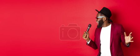 Photo for Passionate black male singer performing against red background, singing into microphone, wearing party outfit, standing over red background. - Royalty Free Image