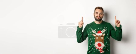 Photo for Winter holidays and christmas. Unamused bearded guy in funny sweater pointing fingers up, showing something unpleasant, white background. - Royalty Free Image