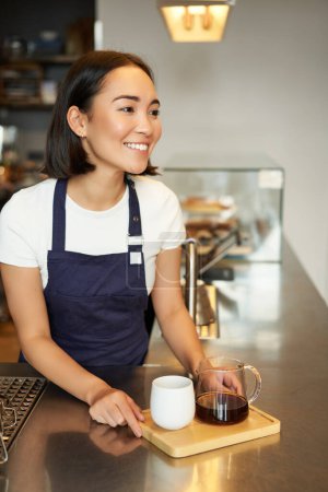 Téléchargez les photos : Plan vertical de fille souriante barista servant du café, fabrication de bière par lots, filtre pour le client dans le café, portant tablier bleu derrière le comptoir. - en image libre de droit