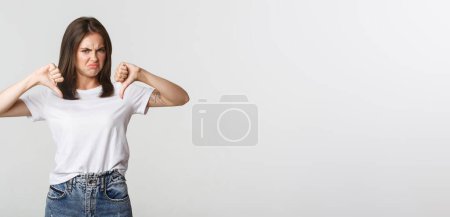 Photo for Disappointed grimacing cute girl showing thumbs-down, express dislike. - Royalty Free Image