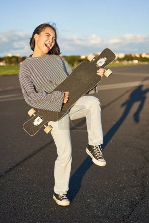 Téléchargez les photos : Drôle asiatique fille appréciant patinage tenant skateboard comme guitare et ombre jouer avoir amusant à l'extérieur. - en image libre de droit