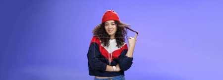 Photo for Stylish and snobbish arrogant curly-haired woman in warm beanie rolling curl on finger smirking and looking with contempt at camera, scorning person as being too cool over blue background. - Royalty Free Image