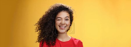 Photo for Lifestyle. Portrait of funny and cool sister with curly hair winking playfully having fun and foolind around showing tongue as playing with siblings adoring spend time with chidren over yellow - Royalty Free Image
