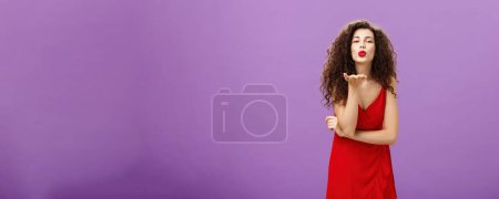 Foto de Mujer enviando un beso apasionado siendo romántica desesperada. de pie en elegante vestido rojo con peinado rizado y maquillaje soplado mwah con sonrisa y labios doblados sosteniendo la palma cerca de la boca sobre la pared púrpura. - Imagen libre de derechos