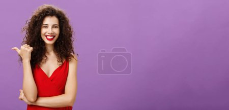 Téléchargez les photos : Je vous suggère de vérifier. Charmante femme européenne élégante joyeuse et amicale avec coiffure bouclée en robe rouge de soirée pointant vers la gauche avec pouce et sourire assuré et confiant montrant l'espace de copie. - en image libre de droit