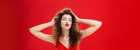 Photo for Waist-up shot of confident flirty and elegant young rich woman. with curly hairstyle in red lipstic and evening dress touching hair folding lips in mwah giving kiss to admirer feeling self-assured. - Royalty Free Image