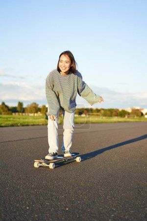 Téléchargez les photos : Mignon asiatique fille équitation skateboard, patinage sur la route et souriant. Skater sur le longboard cruiser profiter en plein air par une journée ensoleillée. - en image libre de droit