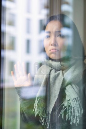 Foto de Asiática mujer se siente mal, se queda en casa, mira fuera de la ventana con celoso, cara angustiada, quiere salir fuera, pero atrapado frío. - Imagen libre de derechos