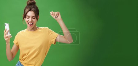 Foto de Mujer feliz fresca y elegante con tatuaje y dientes huecos en camiseta amarilla bailando con alegría escuchando música en auriculares inalámbricos sosteniendo los ojos del teléfono inteligente de cierre y sonriendo de placer sobre la pared verde - Imagen libre de derechos